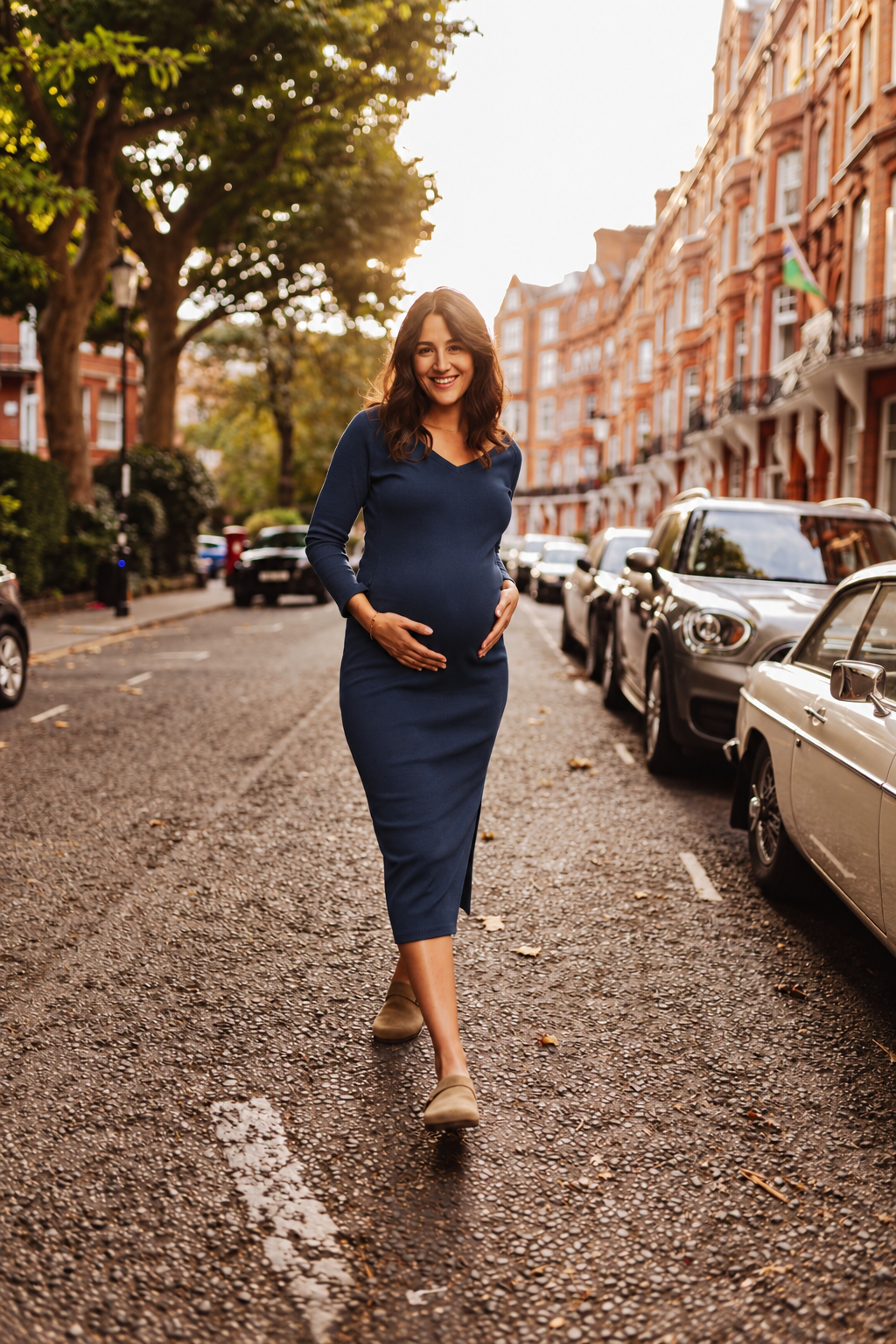 A pregnant woman wearing a navy blue long-sleeve midi dress walks down a sunlit residential street lined with parked cars and trees.